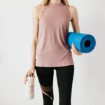 Faceless slim female athlete in sportswear standing with blue fitness mat and water bottle while preparing for indoors workout